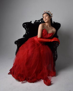 Kırmızı korse, uzun opera eldivenleri ve süslü gotik kraliçe tacı giyen Asyalı güzel bir kadının tam boy portresi. Sandalyede otururken zarif pozlar, stüdyo arka planında izole edilmiş..