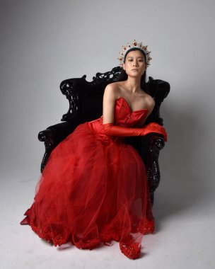 Kırmızı korse, uzun opera eldivenleri ve süslü gotik kraliçe tacı giyen Asyalı güzel bir kadının tam boy portresi. Sandalyede otururken zarif pozlar, stüdyo arka planında izole edilmiş..