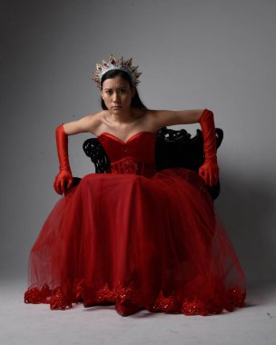 Kırmızı korse, uzun opera eldivenleri ve süslü gotik kraliçe tacı giyen Asyalı güzel bir kadının tam boy portresi. Sandalyede otururken zarif pozlar, stüdyo arka planında izole edilmiş..