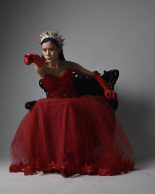 Kırmızı korse, uzun opera eldivenleri ve süslü gotik kraliçe tacı giyen Asyalı güzel bir kadının tam boy portresi. Sandalyede otururken zarif pozlar, stüdyo arka planında izole edilmiş..
