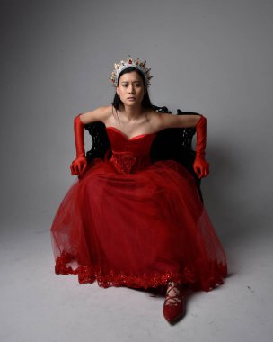 Kırmızı korse, uzun opera eldivenleri ve süslü gotik kraliçe tacı giyen Asyalı güzel bir kadının tam boy portresi. Sandalyede otururken zarif pozlar, stüdyo arka planında izole edilmiş..