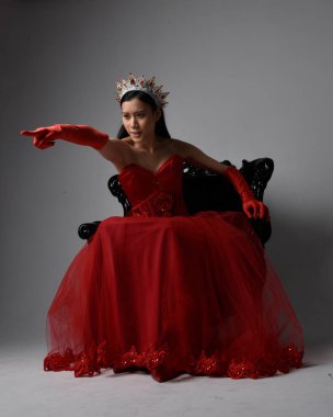 Kırmızı korse, uzun opera eldivenleri ve süslü gotik kraliçe tacı giyen Asyalı güzel bir kadının tam boy portresi. Sandalyede otururken zarif pozlar, stüdyo arka planında izole edilmiş..