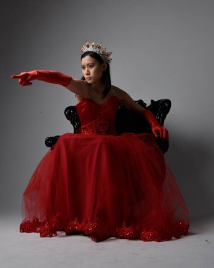 Kırmızı korse, uzun opera eldivenleri ve süslü gotik kraliçe tacı giyen Asyalı güzel bir kadının tam boy portresi. Sandalyede otururken zarif pozlar, stüdyo arka planında izole edilmiş..