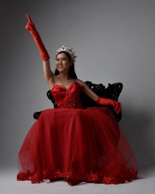 Kırmızı korse, uzun opera eldivenleri ve süslü gotik kraliçe tacı giyen Asyalı güzel bir kadının tam boy portresi. Sandalyede otururken zarif pozlar, stüdyo arka planında izole edilmiş..