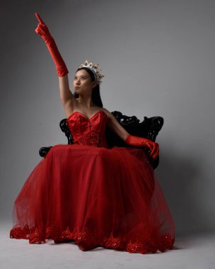 Kırmızı korse, uzun opera eldivenleri ve süslü gotik kraliçe tacı giyen Asyalı güzel bir kadının tam boy portresi. Sandalyede otururken zarif pozlar, stüdyo arka planında izole edilmiş..