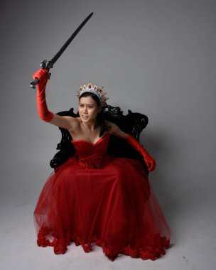Kırmızı korse, uzun opera eldivenleri ve süslü gotik kraliçe tacı giyen Asyalı güzel bir kadının tam boy portresi. Sandalyede otururken zarif pozlar, stüdyo arka planında izole edilmiş..