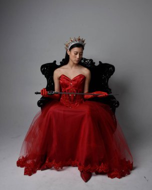 Kırmızı korse, uzun opera eldivenleri ve süslü gotik kraliçe tacı giyen Asyalı güzel bir kadının tam boy portresi. Sandalyede otururken zarif pozlar, stüdyo arka planında izole edilmiş..