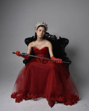 Kırmızı korse, uzun opera eldivenleri ve süslü gotik kraliçe tacı giyen Asyalı güzel bir kadının tam boy portresi. Sandalyede otururken zarif pozlar, stüdyo arka planında izole edilmiş..