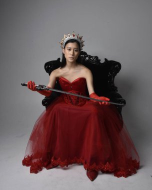 Kırmızı korse, uzun opera eldivenleri ve süslü gotik kraliçe tacı giyen Asyalı güzel bir kadının tam boy portresi. Sandalyede otururken zarif pozlar, stüdyo arka planında izole edilmiş..