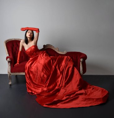 Kırmızı korse, uzun opera eldivenleri ve süslü gotik kraliçe tacı giyen Asyalı güzel bir kadının tam boy portresi. Sandalyede otururken zarif pozlar, stüdyo arka planında izole edilmiş..