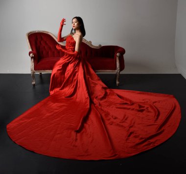 Kırmızı korse, uzun opera eldivenleri ve süslü gotik kraliçe tacı giyen Asyalı güzel bir kadının tam boy portresi. Sandalyede otururken zarif pozlar, stüdyo arka planında izole edilmiş..