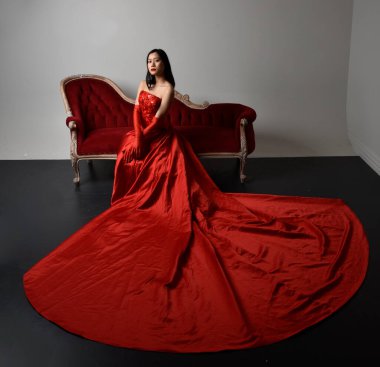 Kırmızı korse, uzun opera eldivenleri ve süslü gotik kraliçe tacı giyen Asyalı güzel bir kadının tam boy portresi. Sandalyede otururken zarif pozlar, stüdyo arka planında izole edilmiş..