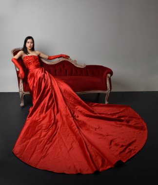 Kırmızı korse, uzun opera eldivenleri ve süslü gotik kraliçe tacı giyen Asyalı güzel bir kadının tam boy portresi. Sandalyede otururken zarif pozlar, stüdyo arka planında izole edilmiş..