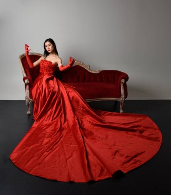 Kırmızı korse, uzun opera eldivenleri ve süslü gotik kraliçe tacı giyen Asyalı güzel bir kadının tam boy portresi. Sandalyede otururken zarif pozlar, stüdyo arka planında izole edilmiş..