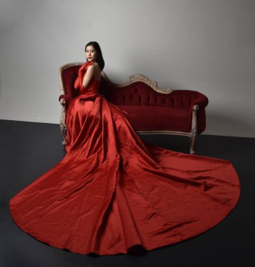 Kırmızı korse, uzun opera eldivenleri ve süslü gotik kraliçe tacı giyen Asyalı güzel bir kadının tam boy portresi. Sandalyede otururken zarif pozlar, stüdyo arka planında izole edilmiş..