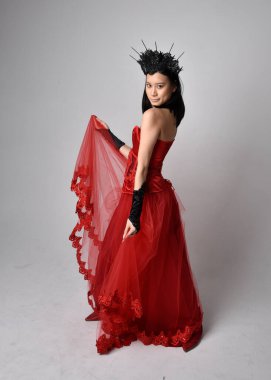 Kırmızı korse takan ve gotik kraliçe tacını takan güzel Asyalı genç bir kadının tam boy portresi. Stüdyonun arka planında zarif duruş.