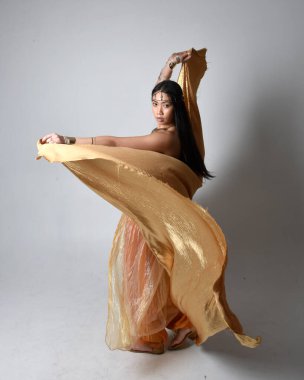 Altın Arap cübbesi giymiş güzel Asyalı bir kadının tam boy portresi. Stüdyo arka planında izole edilmiş, akan kumaşı tutan bir cin gibi duruyor..