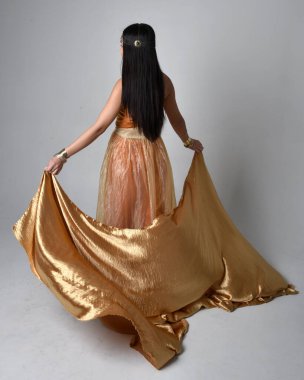 Altın Arap cübbesi giymiş güzel Asyalı bir kadının tam boy portresi. Stüdyo arka planında izole edilmiş, akan kumaşı tutan bir cin gibi duruyor..