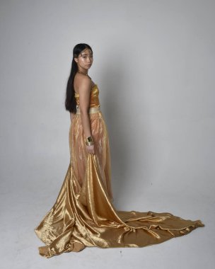 Altın Arap cübbesi giymiş güzel Asyalı bir kadının tam boy portresi. Stüdyo arka planında izole edilmiş, akan kumaşı tutan bir cin gibi duruyor..