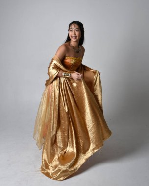 Altın Arap cübbesi giymiş güzel Asyalı bir kadının tam boy portresi. Stüdyo arka planında izole edilmiş, akan kumaşı tutan bir cin gibi duruyor..