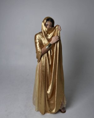Altın Arap cübbesi giymiş güzel Asyalı bir kadının tam boy portresi. Stüdyo arka planında izole edilmiş, akan kumaşı tutan bir cin gibi duruyor..