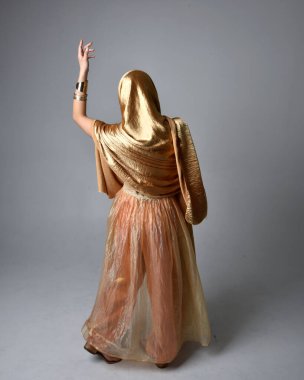 Altın Arap cübbesi giymiş güzel Asyalı bir kadının tam boy portresi. Stüdyo arka planında izole edilmiş, akan kumaşı tutan bir cin gibi duruyor..