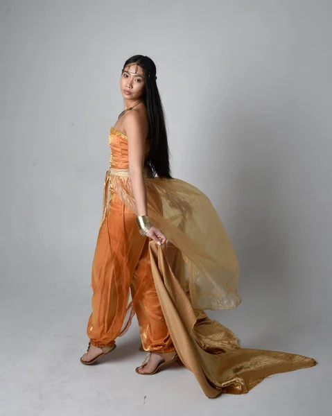 ジーニーのような黄金のアラビア語のローブを身に着けているかなり若いアジアの女性の完全な長さの肖像画 流れるような布を保持立ってポーズ スタジオの背景に孤立 — ストック写真