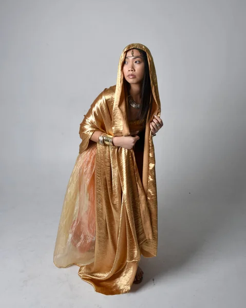 ジーニーのような黄金のアラビア語のローブを身に着けているかなり若いアジアの女性の完全な長さの肖像画 流れるような布を保持立ってポーズ スタジオの背景に孤立 — ストック写真