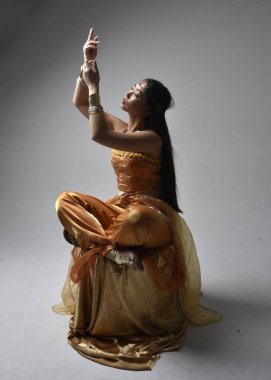 Altın Arap cübbesi giyen, stüdyo arka planında izole bir şekilde oturan, cin gibi uzun boylu Asyalı bir kadının portresi..