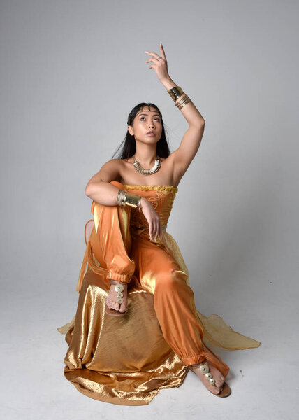 Full length portrait of pretty young asian woman wearing golden Arabian robes like a genie, seated pose, isolated on studio background.