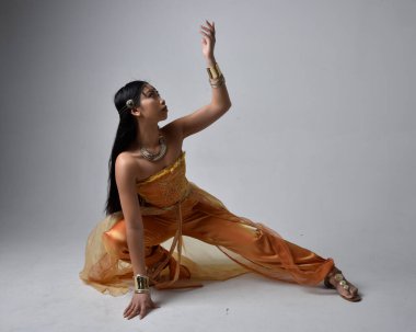 Altın Arap cübbesi giyen, stüdyo arka planında izole bir şekilde oturan, cin gibi uzun boylu Asyalı bir kadının portresi..