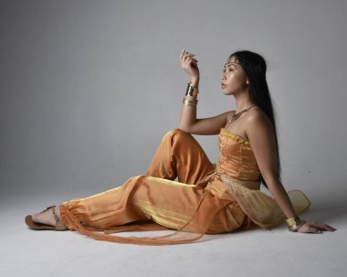 Altın Arap cübbesi giyen, stüdyo arka planında izole bir şekilde oturan, cin gibi uzun boylu Asyalı bir kadının portresi..