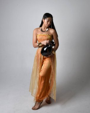 Altın Arap cübbesi giyen, elinde vazo tutan, stüdyo arka planında izole bir vazo tutan Asyalı güzel bir kadının tam boy portresi..