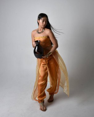 Altın Arap cübbesi giyen, elinde vazo tutan, stüdyo arka planında izole bir vazo tutan Asyalı güzel bir kadının tam boy portresi..