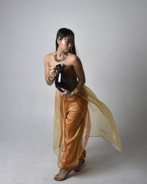 Altın Arap cübbesi giyen, elinde vazo tutan, stüdyo arka planında izole bir vazo tutan Asyalı güzel bir kadının tam boy portresi..