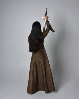 Orta çağ fantezi elbisesi giymiş uzun saçlı güzel Asyalı bir kadının tam boy portresi. Kılıç tutan zarif duruş, stüdyo arka planında izole edilmiş..