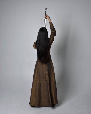 Orta çağ fantezi elbisesi giymiş uzun saçlı güzel Asyalı bir kadının tam boy portresi. Kılıç tutan zarif duruş, stüdyo arka planında izole edilmiş..