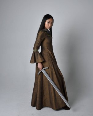 Orta çağ fantezi elbisesi giymiş uzun saçlı güzel Asyalı bir kadının tam boy portresi. Kılıç tutan zarif duruş, stüdyo arka planında izole edilmiş..