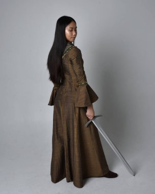 Orta çağ fantezi elbisesi giymiş uzun saçlı güzel Asyalı bir kadının tam boy portresi. Kılıç tutan zarif duruş, stüdyo arka planında izole edilmiş..