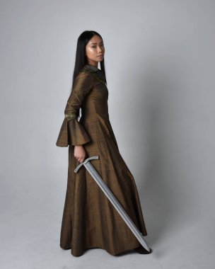 Orta çağ fantezi elbisesi giymiş uzun saçlı güzel Asyalı bir kadının tam boy portresi. Kılıç tutan zarif duruş, stüdyo arka planında izole edilmiş..