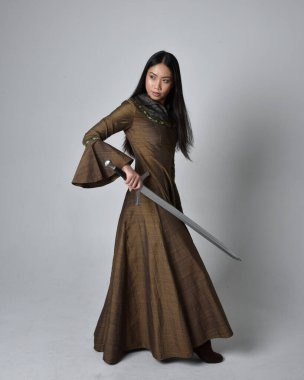 Orta çağ fantezi elbisesi giymiş uzun saçlı güzel Asyalı bir kadının tam boy portresi. Kılıç tutan zarif duruş, stüdyo arka planında izole edilmiş..