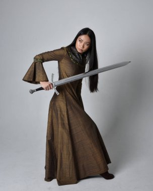 Orta çağ fantezi elbisesi giymiş uzun saçlı güzel Asyalı bir kadının tam boy portresi. Kılıç tutan zarif duruş, stüdyo arka planında izole edilmiş..