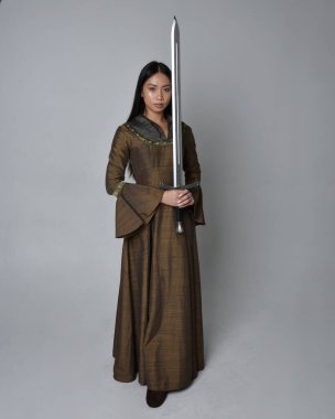 Orta çağ fantezi elbisesi giymiş uzun saçlı güzel Asyalı bir kadının tam boy portresi. Kılıç tutan zarif duruş, stüdyo arka planında izole edilmiş..