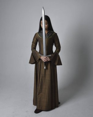 Orta çağ fantezi elbisesi giymiş uzun saçlı güzel Asyalı bir kadının tam boy portresi. Kılıç tutan zarif duruş, stüdyo arka planında izole edilmiş..