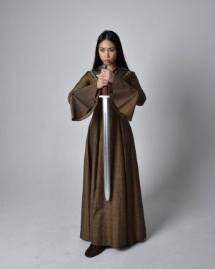 Orta çağ fantezi elbisesi giymiş uzun saçlı güzel Asyalı bir kadının tam boy portresi. Kılıç tutan zarif duruş, stüdyo arka planında izole edilmiş..