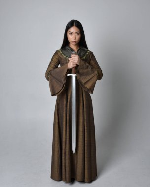 Orta çağ fantezi elbisesi giymiş uzun saçlı güzel Asyalı bir kadının tam boy portresi. Kılıç tutan zarif duruş, stüdyo arka planında izole edilmiş..