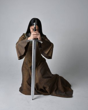 Orta çağ fantezi elbisesi giymiş uzun saçlı güzel Asyalı bir kadının tam boy portresi. Kılıç tutan zarif bir poz, stüdyo arka planında izole edilmiş..