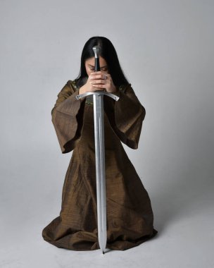 Orta çağ fantezi elbisesi giymiş uzun saçlı güzel Asyalı bir kadının tam boy portresi. Kılıç tutan zarif bir poz, stüdyo arka planında izole edilmiş..