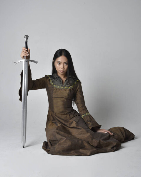 Full length  portrait of beautiful young asian woman with long hair wearing medieval fantasy gown costume. Graceful sitting pose holding a sword,  isolated on studio background.