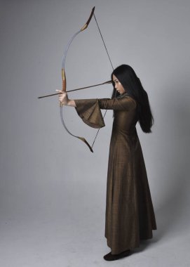 Orta çağ fantezi elbisesi giymiş, uzun saçlı güzel Asyalı bir kadının portresi. Uzun bir yay ve ok tutan zarif bir poz, stüdyo arka planında izole edilmiş..